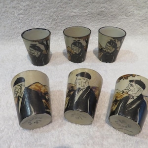 Peut inclure: Six verres à shot en céramique vintage. Chaque verre présente une image peinte à la main d'un vieil homme en costume et chapeau noirs. Les verres ont un fond beige avec des accents marron et noir. Les verres à shot sont disposés sur une surface blanche.