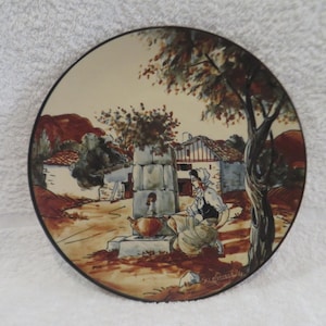 Peut inclure: Assiette décorative ronde avec une scène peinte à la main d'une femme à une fontaine dans un village. L'assiette présente un bord marron et un paysage avec des arbres et des bâtiments dans des tons de marron, beige et vert.