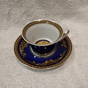 May include: A porcelain teacup and saucer set featuring a deep blue exterior and interior base with intricate gold filigree patterns. The rim of the cup and the edge of the saucer are decorated with a repeating black and gold geometric border design.