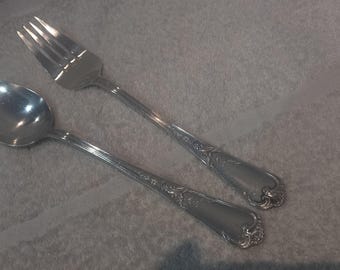 May include: A silver spoon and fork. The spoon has a rounded bowl and a long handle. The fork has four prongs. Both items have decorative detailing on the handles. The set is on a light grey surface.