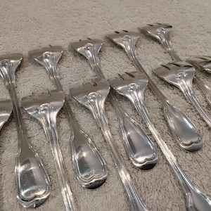 May include: A set of silver serving forks with three prongs. The forks feature an ornate handle design and are laid out on a neutral-toned surface. These forks are likely for serving appetizers or small portions.