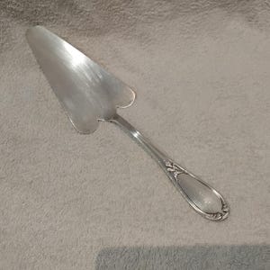 Puede incluir: Una espátula plateada para tartas con una hoja en forma de corazón y un mango decorativo. La espátula está hecha de un metal brillante y reflectante y está diseñada para servir tartas y pasteles. El mango presenta un diseño ornamentado.