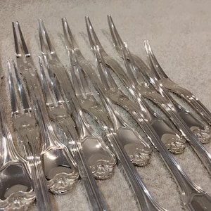 Pode incluir: Uma coleção de garfos de servir prateados com cabos ornamentados e dois dentes. Os garfos estão dispostos em um padrão diagonal, refletindo a luz. A prataria é provavelmente para um ambiente de jantar formal.