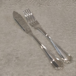 Può includere: Un set di coltello da burro e forchetta argentati. Il coltello ha una lama strutturata e un manico curvo. La forchetta ha quattro rebbi e un manico abbinato. Entrambi i pezzi sono riflettenti e poggiano su una superficie grigio chiaro.