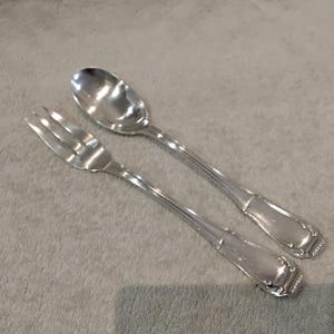 Pode incluir: Um conjunto de garfo e colher prateados. O garfo tem quatro dentes e a colher tem uma tigela arredondada. Ambos os utensílios têm cabos decorativos com um design retangular e um pequeno detalhe padronizado na extremidade. O conjunto está sobre uma superfície cinzenta.