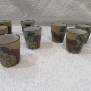 Peut inclure: Ensemble de huit petits verres à shot en céramique. Chaque verre présente un fond beige avec une illustration marron et noire d'une personne. Les verres ont un intérieur blanc et un bord marron.