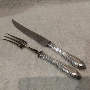 May include: A silver-toned carving knife and a matching two-pronged carving fork. Both utensils feature ornate, embossed handles with intricate detailing and a polished metallic finish, paired with smooth, tapered blades and tines.