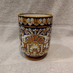 Peut inclure: Un vase en céramique avec un design détaillé. Le vase présente un fond bleu avec des accents jaunes et blancs. Le motif comprend des chérubins et un motif de coquillage. La base et le bord sont jaunes.