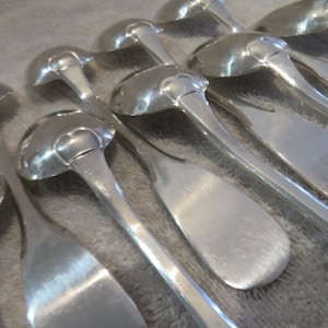 Peut inclure: Un ensemble de cuillères en argent avec des manches ornés. Les cuillères sont disposées en rangée, les manches pointant vers le spectateur. Les cuillères sont toutes polies et brillantes.