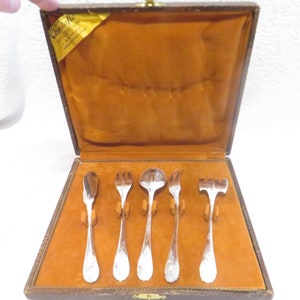 May include: A set of six silver-plated serving utensils in a brown leatherette case. The utensils include a butter knife, a fork, a spoon, a serving spoon, a fork, and a small hammer.