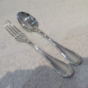 Könnte beinhalten: Ein silbernes Löffel- und Gabel-Set. Der Löffel hat eine abgerundete Schale, und die Gabel hat vier Zinken. Beide Utensilien haben dekorative Details an den Griffen. Das Set besteht aus einem glänzenden, reflektierenden Metall.