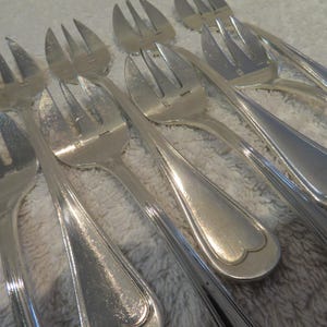 May include: A collection of silver-colored forks with three tines each. The forks have a classic design with a rounded handle and a decorative edge. The silverware is arranged on a light-colored surface.