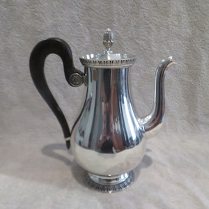 May include: A silver coffee pot with a black wooden handle and a decorative lid. The pot has a round body and a flared base.