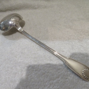 Puede incluir: Un cucharón de plata con un cuenco redondeado y un mango largo y delgado. El mango tiene un diseño decorativo en el extremo.