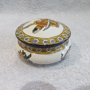 Peut inclure: Une boîte à bijoux en céramique blanche avec un couvercle rond. Le couvercle est décoré d'un motif jaune, orange et bleu d'une épée et d'un bouclier. La base de la boîte est décorée d'un motif floral jaune, vert et bleu.