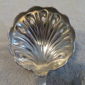 May include: A silver serving spoon with a decorative shell design and holes for straining. The spoon is made of polished metal and has a long handle.