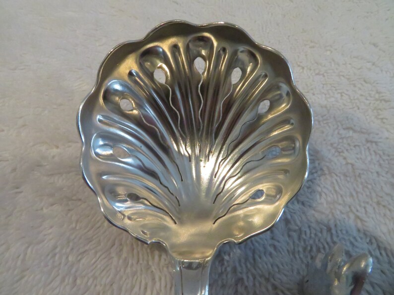 May include: A silver serving spoon with a decorative shell design and holes for straining. The spoon is made of polished metal and has a long handle.