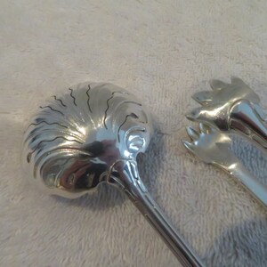 May include: A set of two silver serving utensils. One utensil is a spoon with a decorative, scalloped design. The other utensil is a pair of tongs with a claw-like design.