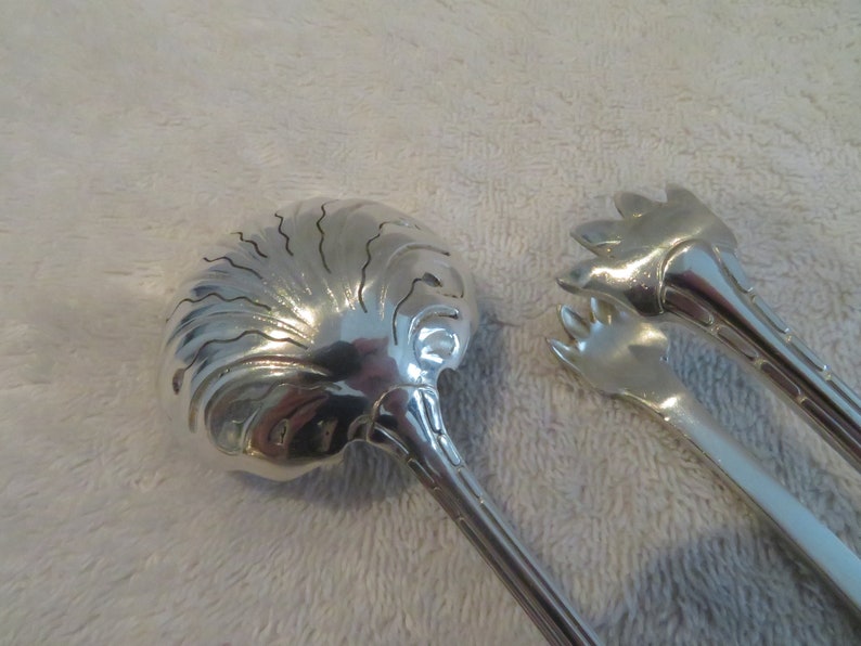 May include: A set of two silver serving utensils. One utensil is a spoon with a decorative, scalloped design. The other utensil is a pair of tongs with a claw-like design.