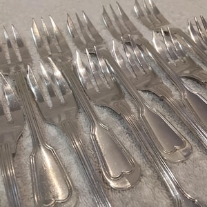 Könnte beinhalten: Eine Sammlung von Silber-Gabeln mit klassischem Design. Die Gabeln haben vier Zinken und einen dekorativen Griff mit herzförmigem Detail. Das Besteck ist auf einer hellen Oberfläche angeordnet, die das Licht reflektiert.