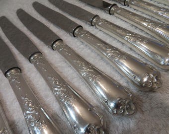 May include: A set of silver dinner knives with ornate handles. The knives have a polished finish and are arranged in a row.