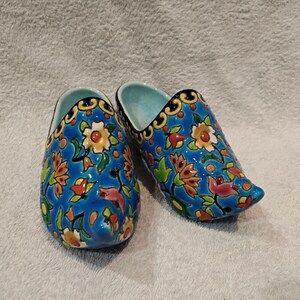 Könnte beinhalten: Ein Paar Keramik-Holzschuhe, jeweils bemalt mit einem leuchtend blauen Hintergrund und bunten Blumenmustern. Die Holzschuhe haben eine hellblaue Innenseite und einen dekorativen Goldbesatz.