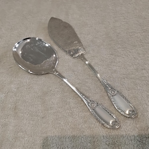 May include: A silver serving spoon and butter knife set. The spoon has a rounded bowl, and the knife has a wide, flat blade. Both utensils feature ornate, detailed handle designs with a central oval accent.