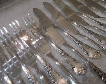 May include: A set of silver forks and knives with ornate handles. The silverware is arranged in a row, with the forks on the left and the knives on the right.