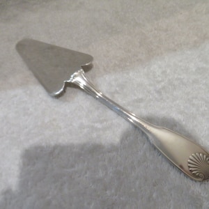 Peut inclure: Une pelle à gâteau argentée avec un bord festonné et un motif de coquillage sur la poignée. La pelle a une lame triangulaire et une longue poignée fine. Ustensile de service classique.