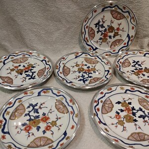Op de afbeelding: Een set van zes witte porseleinen borden met een bloemenontwerp. De borden hebben een zilveren rand en een centrale opstelling van oranje, gouden en blauwe bloemen met bruine en blauwe accenten. De borden zijn ovaal.