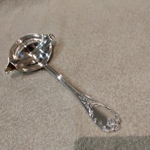 May include: A silver-colored, ornate ladle with a spout and a decorative handle. The ladle has a rounded bowl with a small spout for pouring. The handle features an engraved floral design.