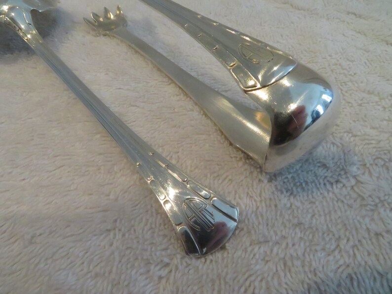 May include: A pair of silver-plated ice tongs with an ornate design. The tongs have a decorative handle with a monogram.