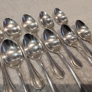 Könnte beinhalten: Eine Sammlung glänzender, silberner Löffel, die auf einer hellen Oberfläche angeordnet sind. Die Löffel haben ein klassisches Design mit länglichen Griffen und ovalen Schalen, die das Umgebungslicht reflektieren. Die Löffel sind wahrscheinlich zum Servieren oder Essen.
