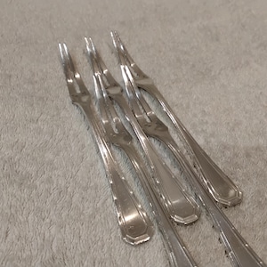 Pode incluir: Quatro garfos prateados com dois dentes cada, dispostos diagonalmente sobre uma superfície cinza claro texturizada. Os garfos têm um padrão decorativo nas alças e são feitos de metal brilhante e refletivo.