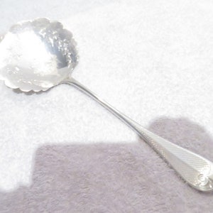 Peut inclure: Une cuillère de service en argent avec un bord festonné et un manche orné. La cuillère est sur un fond blanc.