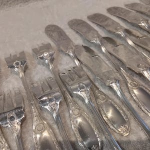 May include: An assortment of silver-colored flatware, including forks and knives, arranged on a light-colored surface. The silverware has decorative detailing on the handles and a shiny finish, suggesting a formal dining set.