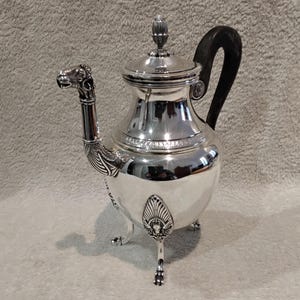 Peut inclure: Une tasse en céramique blanche avec une anse noire et le texte "But First, Coffee" en gras noir. La tasse est posée sur un fond flou, suggérant un cadre chaleureux.