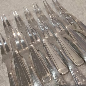 May include: A set of silver-toned metal cocktail forks, each featuring two tines and a handle decorated with a series of etched horizontal lines. The forks are arranged in a parallel formation, showcasing the reflective metallic finish and the uniform design of the cutlery pieces.