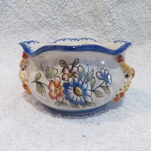 Peut inclure: Un bol en céramique blanche avec des motifs floraux bleus et jaunes et des bords festonnés. Le bol a deux anses en forme de fleurs stylisées.