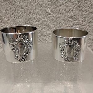 Könnte beinhalten: Zwei silberfarbene Serviettenringe aus Metall mit einer polierten, reflektierenden Oberfläche. Jeder Ring ist auf der Vorderseite mit einem markanten, erhabenen Wappen oder schildförmigen Emblem versehen, das durch filigrane Schnörkel besticht.