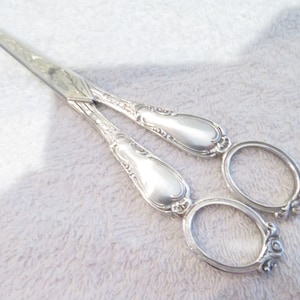 Puede incluir: Tijeras plateadas con mangos ornamentados y detalles florales. Las tijeras tienen una hoja puntiaguda y están cerradas.