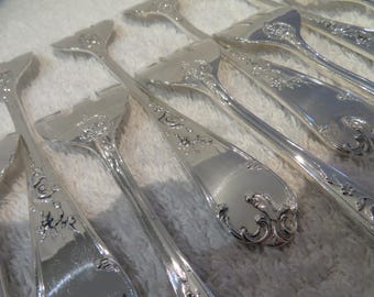 Op de afbeelding: Close-up van verschillende stukken sierlijk zilver bestek. Het bestek omvat vorken, opscheplepels en ander keukengerei met gedetailleerde bloemen- en krulontwerpen. De stukken zijn gerangschikt op een wit, getextureerd oppervlak.