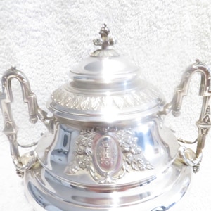 Puede incluir: Una tetera plateada y ornamentada con tapa y dos asas. La tetera presenta detalles decorativos florales y de escudo. La tapa está rematada con un pequeño adorno. La tetera es probablemente de metal.