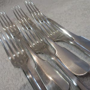 Pode incluir: Um conjunto de seis garfos de prata com cabos ornamentados. Os garfos estão dispostos em forma de leque, com os dentes apontando para cima.
