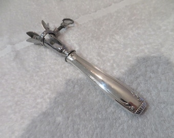 May include: A silver-colored wine bottle opener with a decorative handle. The opener has a lever mechanism and a ring at the top. The handle has an ornate design at the base. The opener is on a white surface.