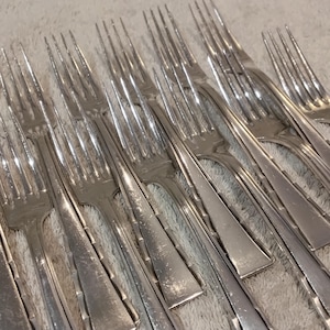 Pode incluir: Uma coleção de garfos prateados com dentes, dispostos sobre uma superfície de cor clara. Os garfos têm um design clássico com um cabo longo e quatro dentes. Os talheres são provavelmente para jantar.