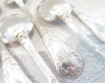 May include: Close-up of several silver serving spoons with ornate floral and scrollwork detailing. The spoons have a reflective, polished surface and feature a monogram on the handle. The spoons are arranged on a light-colored surface.
