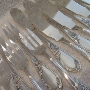 May include: A set of silver-plated forks and knives with ornate handles. The handles have a floral design and a white enamel inlay. The forks and knives are arranged in a row on a white surface.