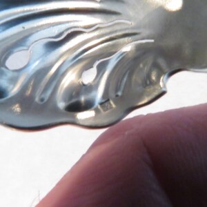 May include: A silver metal leaf-shaped object with two holes in it. The object is made of a thin metal and has a wavy design.