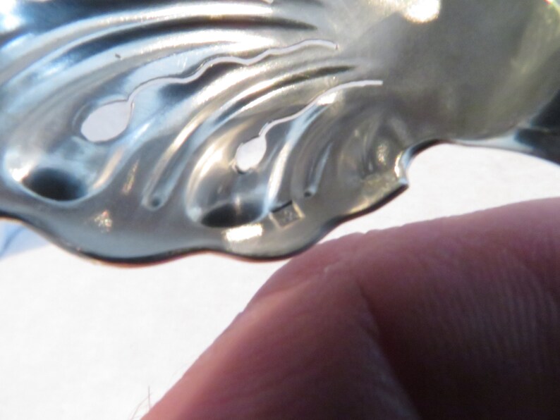 May include: A silver metal leaf-shaped object with two holes in it. The object is made of a thin metal and has a wavy design.